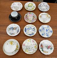 Photo showing 12 teacups and saucers of various floral designs arranged on a wooden surface. Includes black cup with rhinestone handle and multiple floral patterns in vibrant colors.