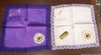Two handkerchiefs side by side on table, one deep purple and one pink with lace borders, brass lighter atop pink one.