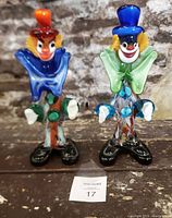 Two colorful Murano glass clown figurines side by side on a wooden surface with stone wall background.