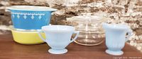 Wide shot showing four covered Pyrex bowls including blue garland bowl, yellow bowl, clear glass bowl with lid, plus Delphite blue creamer and sugar set in front.