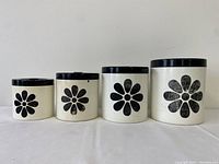 Front view of four black and white canisters with white daisy design in ascending size order.