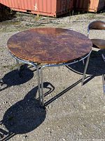 Round Mid-Century Modern dining table with wood veneer top and chrome metal base, shown outdoors on gravel surface.