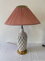 Full view of the Mid-Century Modern style white ceramic table lamp with textured wavy pattern and gold metal base, topped with a pink pleated fabric lamp shade with brass trim.