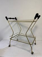 Side view of metal and glass bar cart showing the gold frame, removable bottom glass shelf, black grips on handles and caster wheels.
