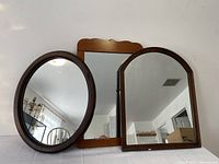 Three solid wood framed mirrors next to each other on a flat surface showing the overall style and shape: oval, rectangular scalloped pine and arched mirror.