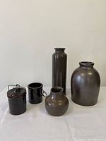 Five pieces of brown pottery with varied shapes and sizes placed against a neutral background. Includes two lidded jars with metal clasps, a small rounded vase, a tall cylindrical vase, and a large rounded jar. Surfaces show wear and imperfections.