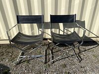 Pair of black leather directors chairs with chrome frames photographed outdoors on gravel in front of a light-colored corrugated metal wall.