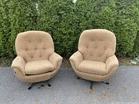 Pair of beige mid-century modern style swivel egg chairs outdoors on pavement with evergreen shrub background. Shows both chairs side by side.