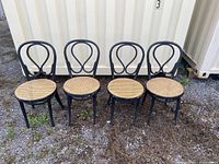 Four bentwood chairs with cane seats painted black and showing wear, placed outdoors in front of a white container.