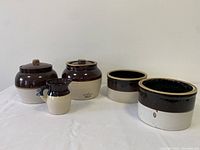 Five ceramic pottery bean pots lined up, showing two with lids and varying sizes and shapes, two-tone brown and beige glaze.