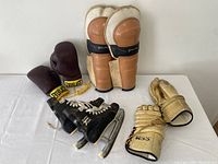 Lot of vintage sports gear including boxing gloves, hockey gloves, shin guards, and ice skates on a white surface.