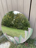 Full view of the circular mirror leaning against an exterior wall showing reflection of trees and sky, highlighting the decorative colored glass detailing at the bottom edge.