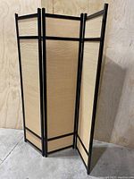 View of the three-panel room divider screen partially folded, showing the black wooden frame and natural woven fibre panels