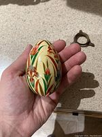Hand holding wooden egg with embossed enamel floral decoration, showing size scale and detail.