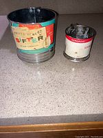 Two antique metal flour sifters side by side on countertop showing labels and condition.
