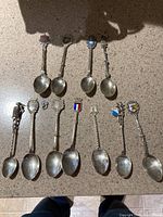 Top down photo of 9 silver spoons arranged in two rows, featuring crests, enamel flags, and ornate handles.
