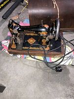 Antique black Singer sewing machine with gold decoration on wooden base, showing wear and connected to electrical cord.
