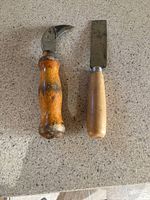 Photo showing two antique hand tools, one with a curved blade and wooden handle, the other with a straight flat blade and wooden handle on a beige surface.