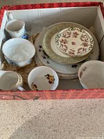 Box containing assorted small plates, saucers, and cups with floral patterns and gold trim.