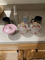 Photo showing all items together: two black ceramic mugs, clear glass Pepsi bottle, clear glass mug with blue emblem, two decorative covered glass bowls, and pink ceramic sugar bowl with lid.