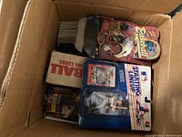 Box containing several baseball card packs, collectible figures, and trading card boxes