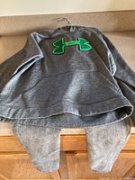 Grey Under Armour hoodie folded with front green logo visible and grey jeans underneath on surface.