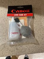 Canon Lens Care Kit with cleaning solution, blower, and brush in clear plastic packaging.