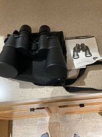 Full view of the black Emerson binoculars with lens covers, black soft carrying pouch, and instruction manual on a beige countertop.