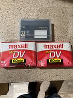 Photo showing two sealed Maxell Mini DV 60-minute tapes and one used Panasonic Mini DV tape case on a table surface.