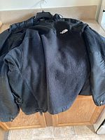 Black North Face fleece jacket with full zipper, laid open over counter, showing front and logo on left chest.