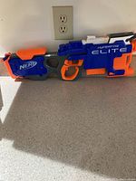Photo of a Nerf Hyperfire Elite blaster in blue, orange, and gray on a countertop with an electrical outlet in the background.