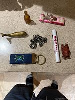 Overview of the full assortment on a counter showing all seven items including figurine, pen, keychains, chain, brass fish, and small bottle.