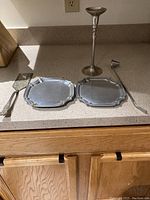 Photo showing six assorted pewter and nickel items including two rectangular serving platters, one candlestick holder, one candle snuffer, a cake server, and a long-handled utensil.