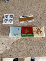 Lot contents on countertop: block of four U.S. stamps featuring man's profile, Goldmark 23K gold leaf box, and three vintage matchbooks with holiday/peace themes.