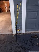 Yellow fishing rod with black handle and attached silver spinning reel leaning against a wall near a garage door on an asphalt driveway.