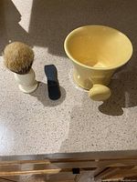 Shaving brush, black razor strop, and yellow ceramic mug on countertop seen from above