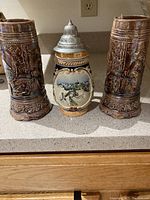 Photo showing one ceramic beer stein and two tall brown ceramic mugs with raised relief design on the exterior.
