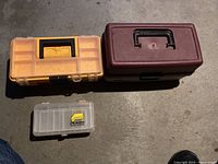 Three fishing tackle boxes placed side by side on a concrete floor: an orange/clear box, a larger maroon box with handle, and a small clear box with yellow Plano logo.