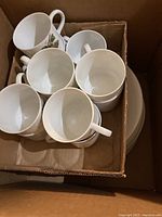 Box containing multiple white porcelain cups, stacked together, showing handles and inside surfaces.