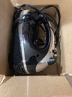 Black and silver Vonshelf hand mixer from side view in a cardboard box showing the main unit with power cord neatly coiled.