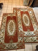 Two smaller rugs measuring approximately 52x27 inches each laid on tile floor showing orange, cream, green, and brown floral medallion pattern with scrollwork borders.
