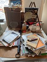Overall view of various art supplies on a table including calligraphy pads, paper cutter, wooden easel and paints.
