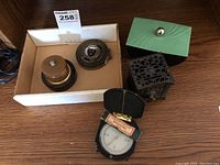 Forestry Compass in open leather case, metal measuring tape, ink well holder with marble base, metal piggy bank, and green dresser jar on wood surface