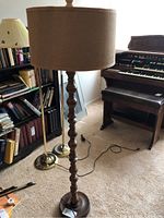 Full view of vintage floor lamp with tall turned wood base and burlap-look drum shade standing on carpeted floor next to bookshelf and organ.