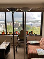Two tall floor lamps standing side-by-side by a window with a city view, visible in room with table and wheelchair.