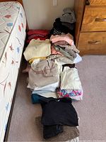 Full view of assorted stacked ladies clothing including sweaters, patterned blouses, and knit tops laid on bedroom carpet next to a bed and dresser.