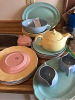 Various pastel colored dishes including platters, serving bowls, plates, a yellow teapot, small creamers, and a sugar bowl arranged on kitchen counter.