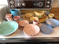Full set arranged on stove showing pink, yellow, blue, and green ceramic pieces including cups, bowls, platters, and sauce boat.