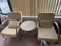Set of two vintage metal frame rocking chairs with beige floral cushions, a round side table with matching patterned tablecloth between them placed near window