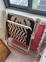 Two folded chairs side by side; one is a folding chair with red and white striped vinyl and the other is a woven lounger chair with black and cream geometric pattern.